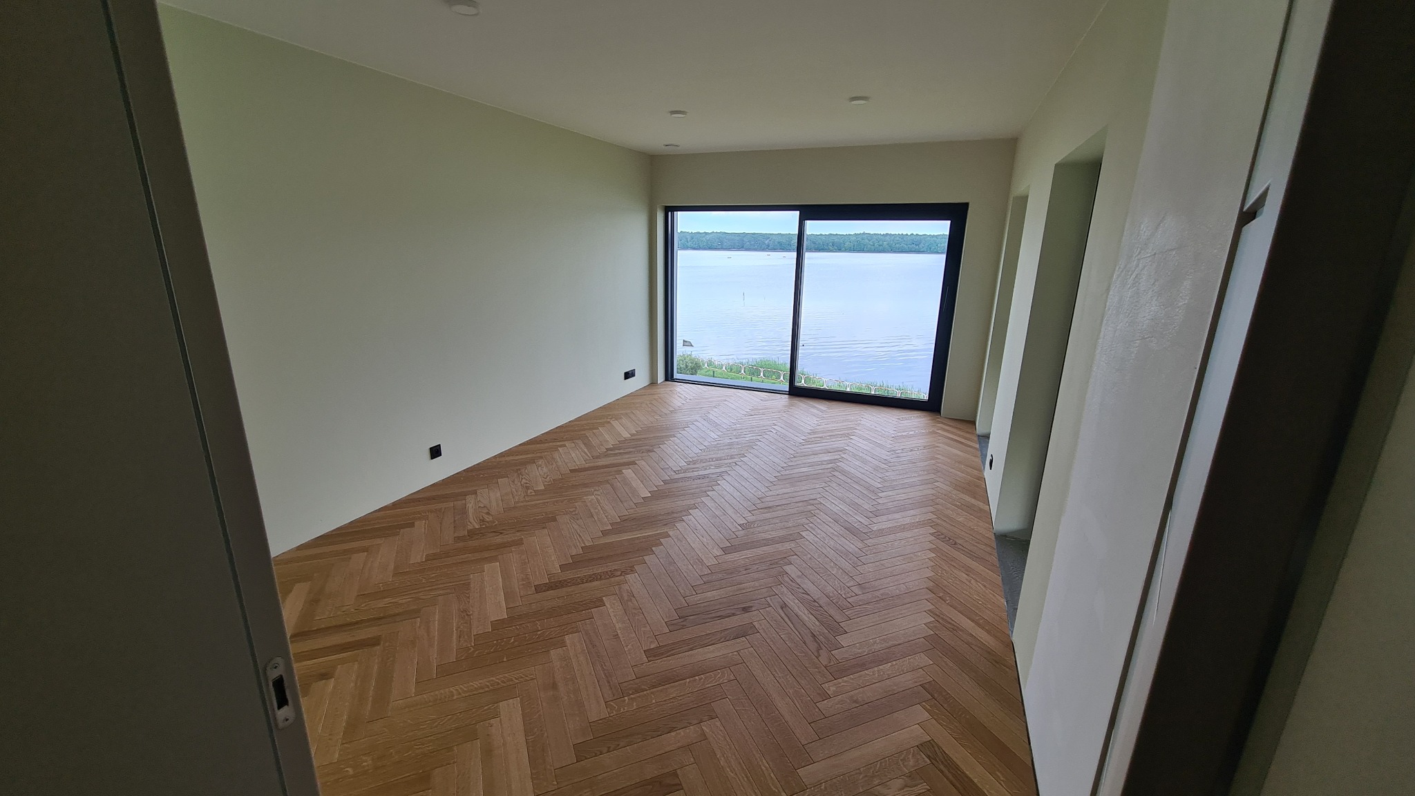 Jasne wnętrze z parkietem w jodełkę, duże okno z widokiem na jezioro. Minimalistyczny design, świeżo po remoncie, widok na zewnątrz przez przesuwne okno.