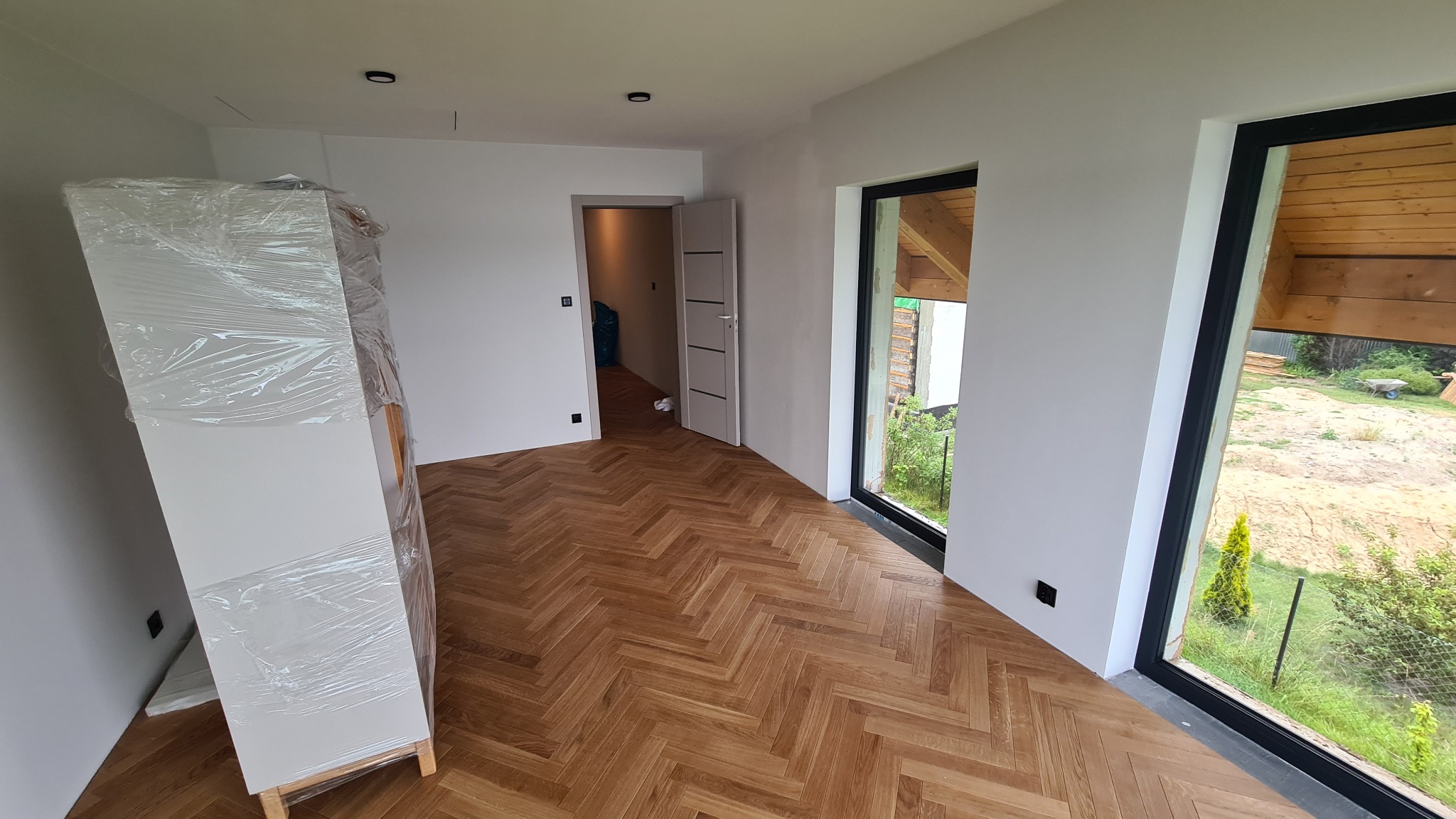 Jasne wnętrze z parkietem w jodełkę, duże okna z czarnymi ramami i drzwi wewnętrzne. Szafa w folii ochronnej. Minimalistyczny design, dobrze doświetlone.