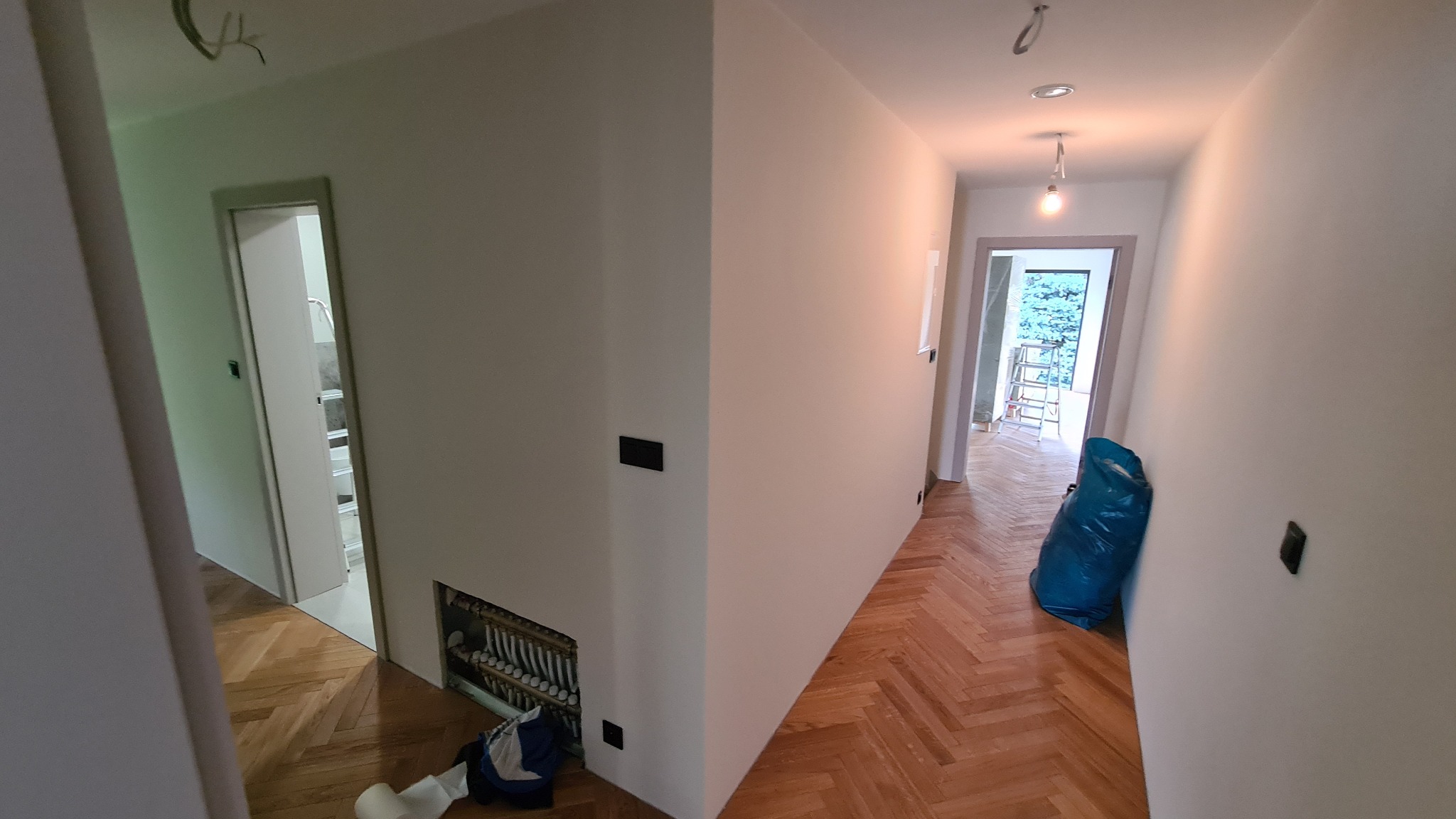 Wnętrze po malowaniu: białe ściany, parkiet jodełka, widoczne gniazdka elektryczne, otwarte drzwi. Torba budowlana w rogu. Widok na schody przez drzwi.