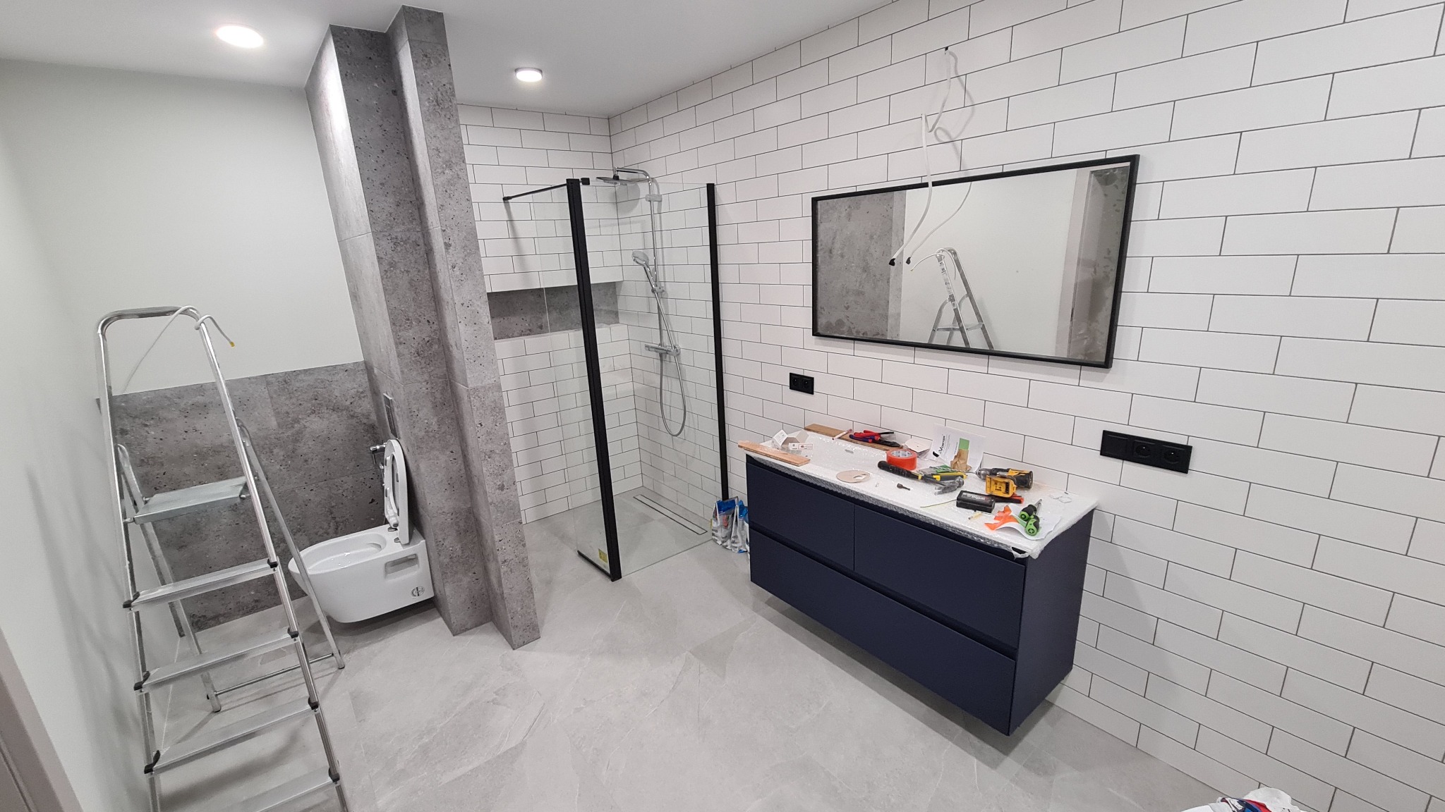 Nowoczesna łazienka w trakcie remontu: białe płytki, kabina prysznicowa z czarnymi ramami, umywalka z szafką w kolorze granatowym. Widoczne narzędzia i drabina.