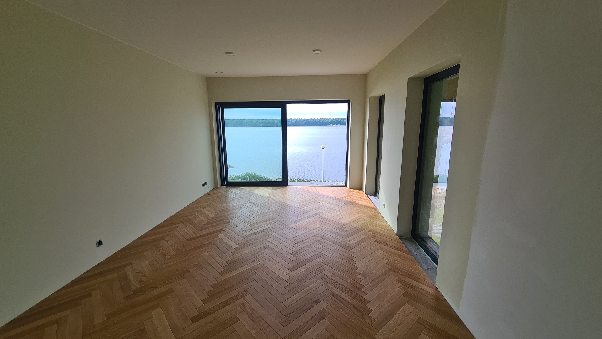 Jasne wnętrze z parkietem ułożonym w jodełkę, duże okno z widokiem na jezioro. Świeżo pomalowane ściany w odcieniach bieli i beżu, widoczne gniazdka elektryczne.