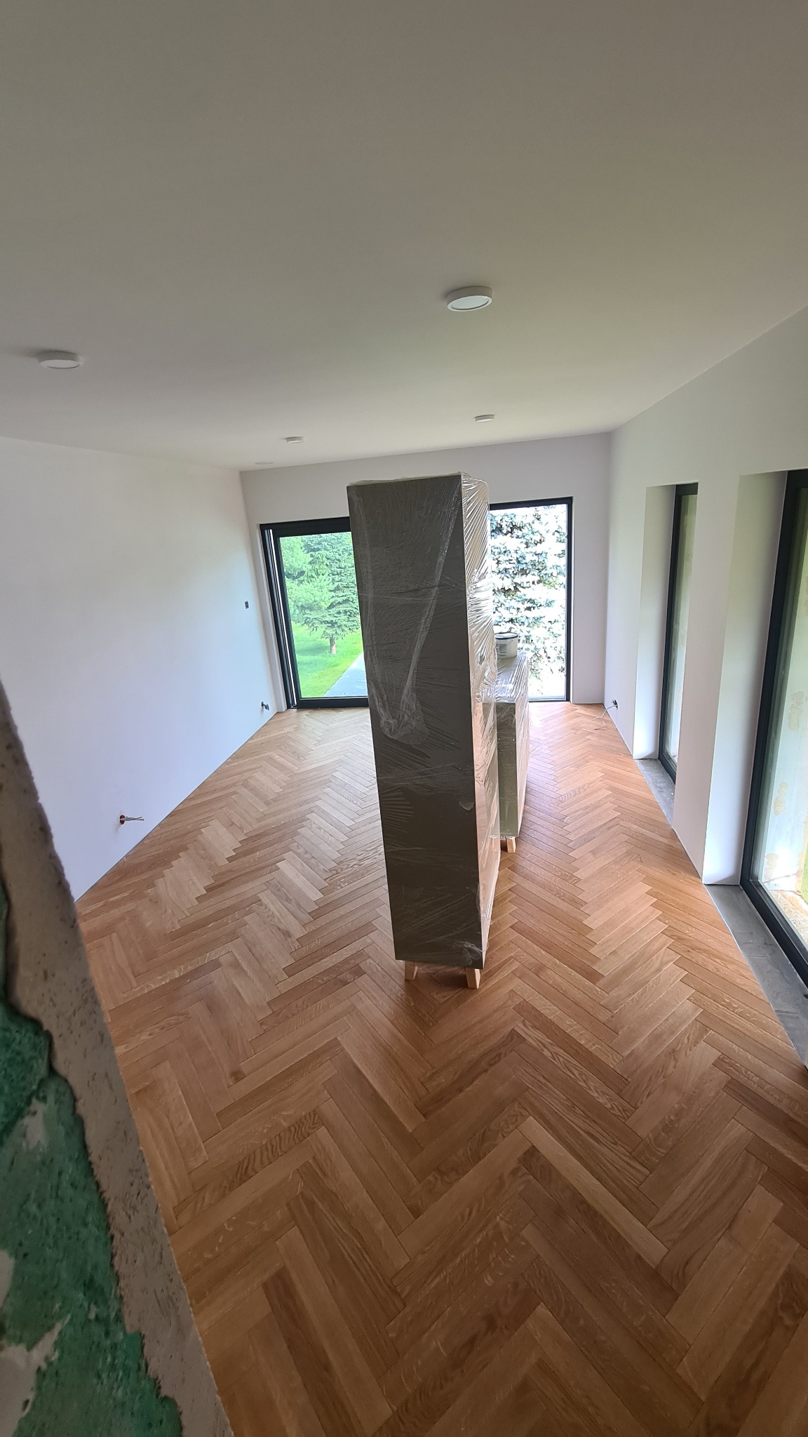 Nowoczesne wnętrze z parkietem w jodełkę, duże okna z widokiem na zieleń. Meble zabezpieczone folią. Częściowo wykończone ściany. Minimalistyczny design.