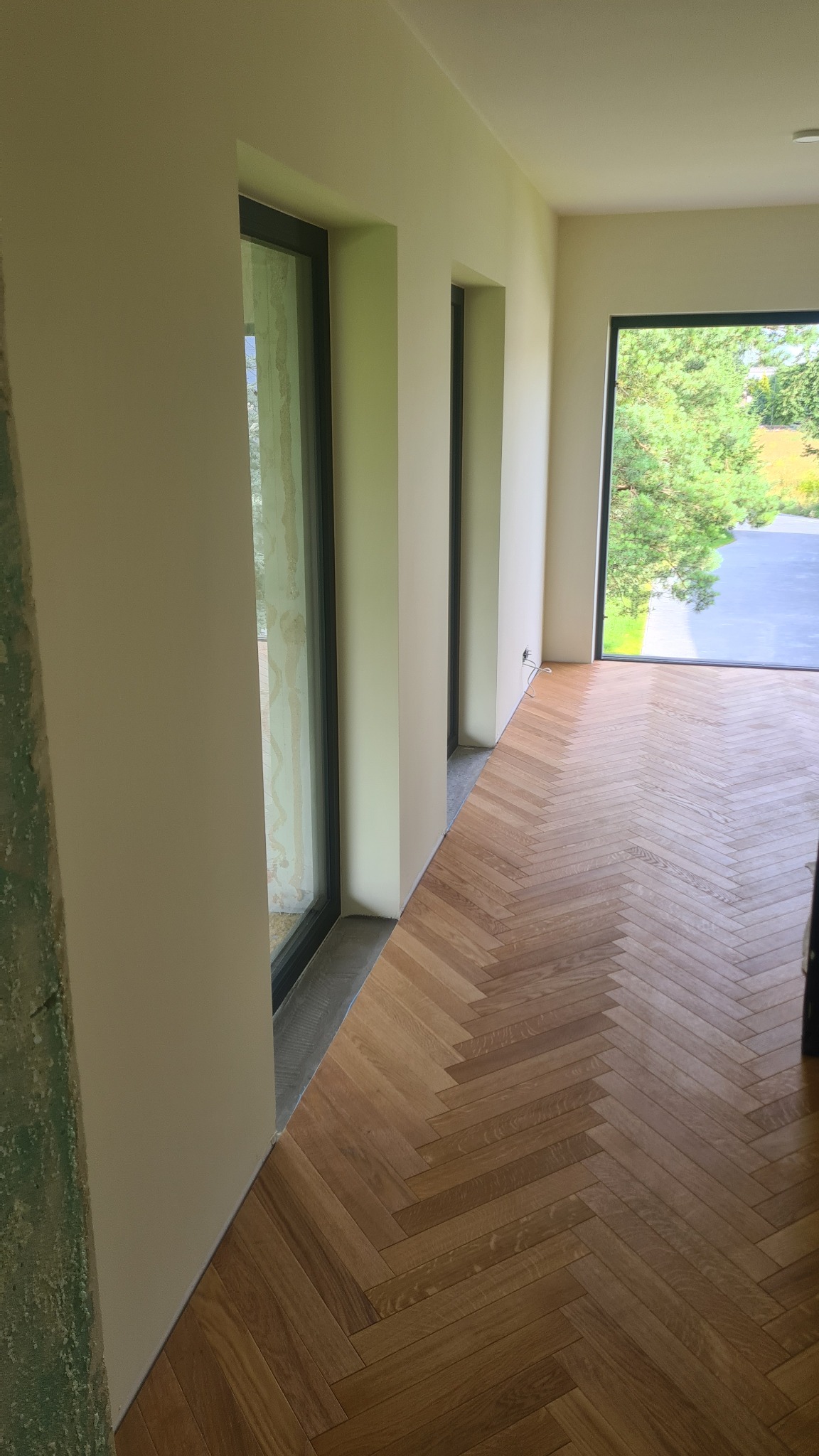 Ułożony parkiet w jodełkę w jasnym wnętrzu z oknami w Lublinie. Widoczne fragmenty ścian i okien, częściowo wykończone, w trakcie prac wykończeniowych.