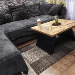 Dębowy stolik kawowy z czarnymi, stalowymi nogami stoi na wzorzystym dywanie obok szarej, narożnej sofy. Na blacie stolika stoi latte macchiato, szklany słoik z rośliną i rozsypane ziarna kawy.