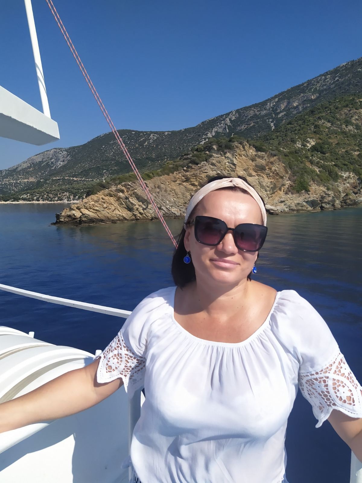 Kobieta w okularach przeciwsłonecznych na pokładzie łodzi z widokiem na górzysty brzeg morski w słoneczny dzień.