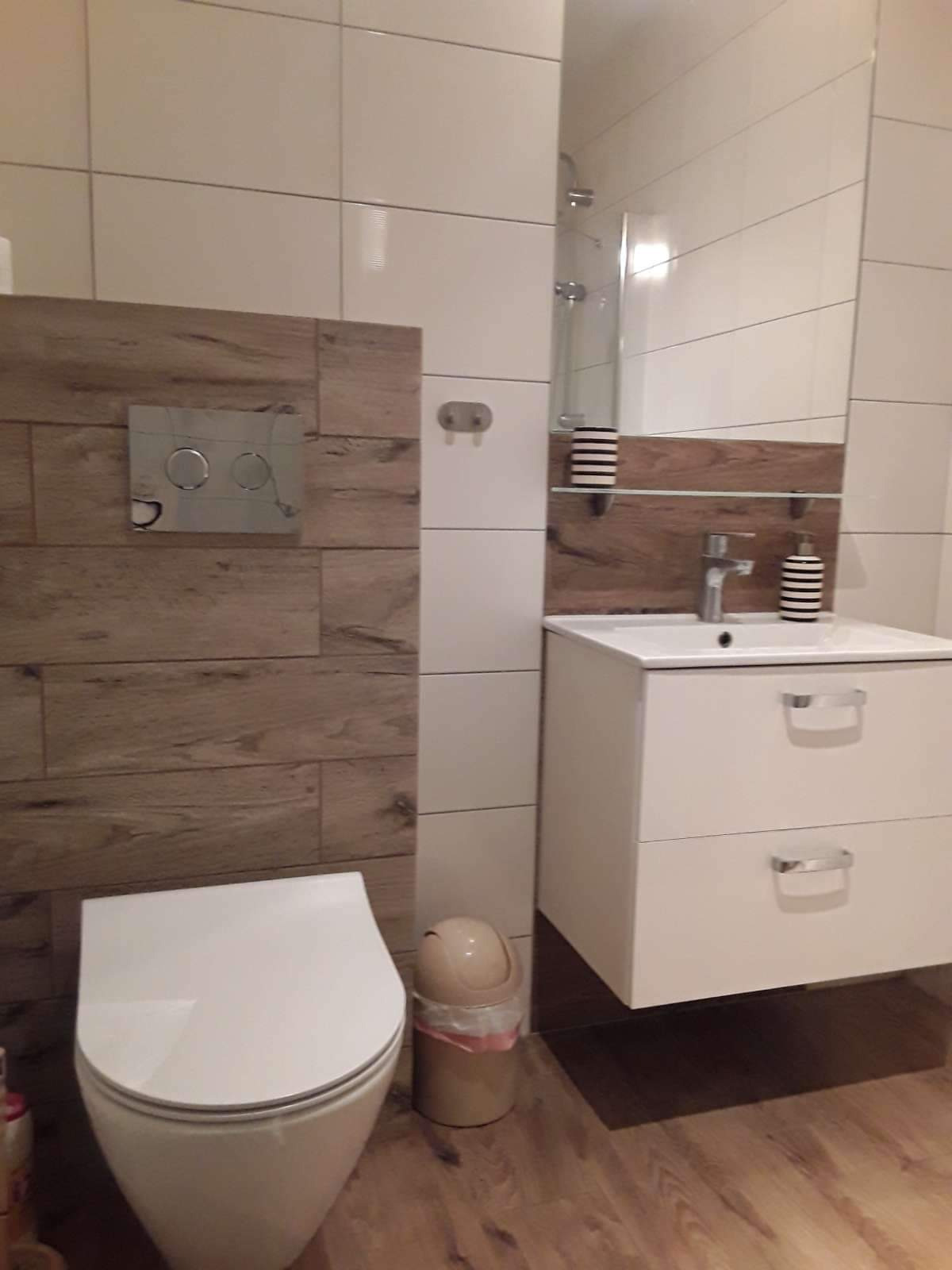 Nowoczesna łazienka z białymi płytkami, drewnopodobną ścianką, podwieszaną toaletą i szafką z umywalką, uzupełniona o drewnianą półkę i akcesoria w paski.