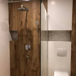 Nowoczesna łazienka z kabiną prysznicową walk-in, drewnopodobne płytki na ścianach i podłodze w strefie prysznica, białe płytki w pozostałej części, podtynkowa toaleta z przyciskiem dwudzielnym...