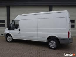 Biały samochód dostawczy Ford Transit na tle szarej, metalowej ściany garażu. Widoczny z boku, lekko od tyłu. Koła lekko zabrudzone.