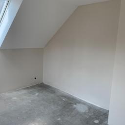 Grafixbud Ilya Karshunou - Pomalowane na beżowo poddasze ze skośnym oknem dachowym i podłogą z resztkami farby, widoczny fragment gniazdka elektrycznego.