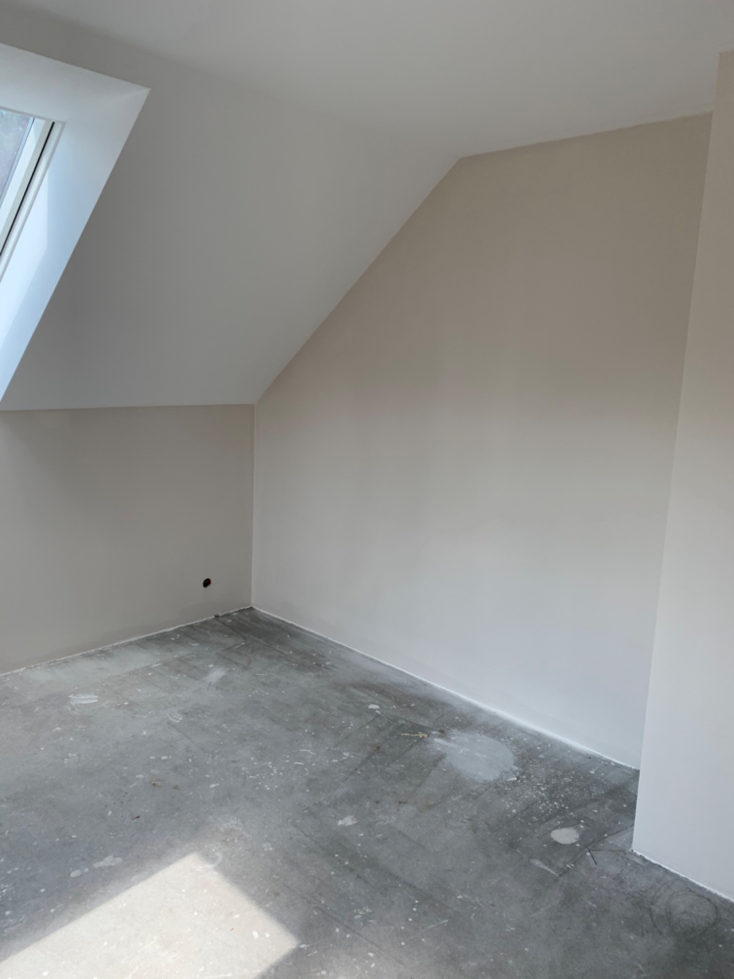 Pomalowane na beżowo poddasze ze skośnym oknem dachowym i podłogą z resztkami farby, widoczny fragment gniazdka elektrycznego.