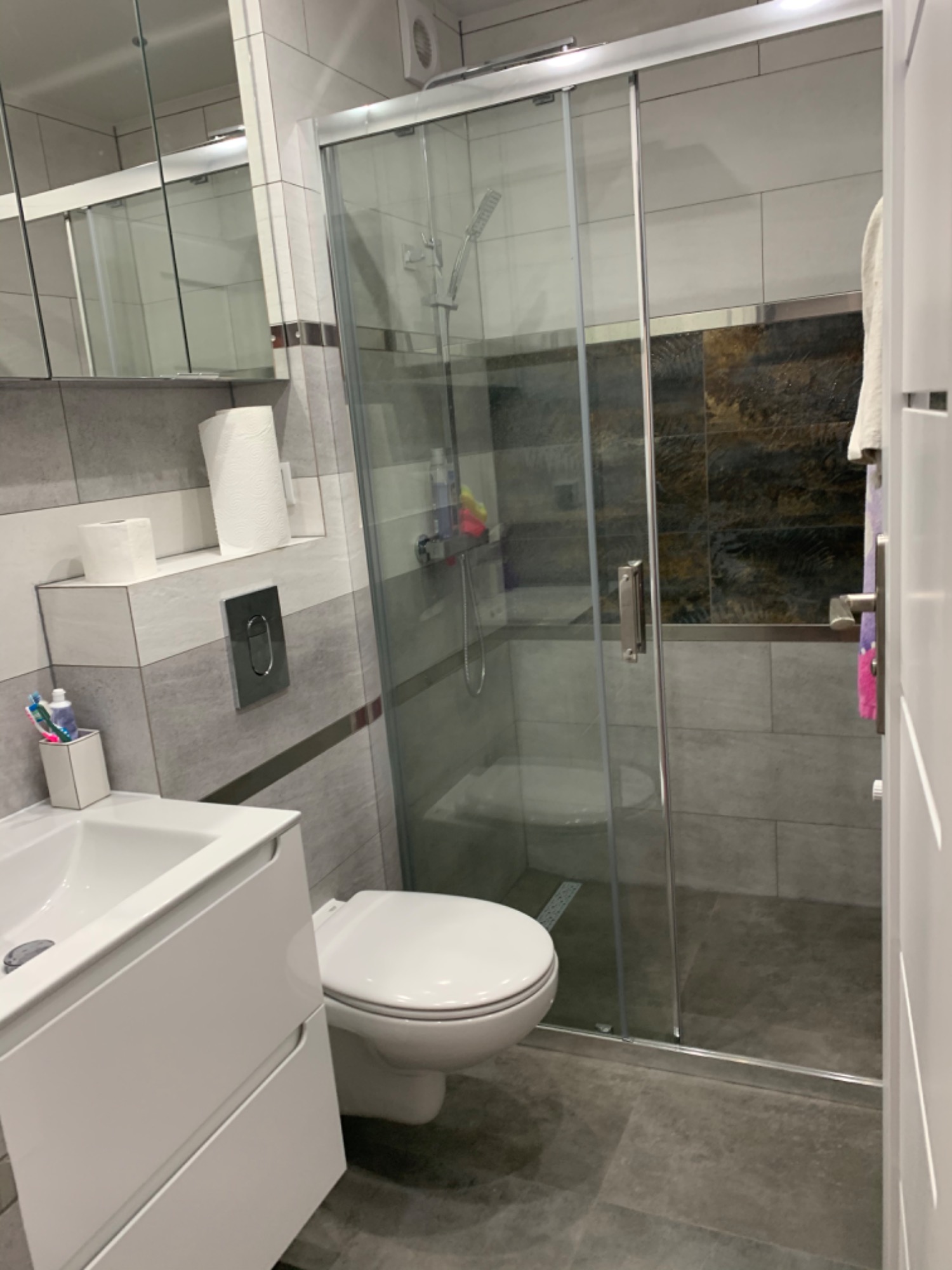 Nowoczesna łazienka z kabiną prysznicową ze szklanymi drzwiami przesuwnymi, umywalką z szafką w kolorze białym, wiszącą toaletą i szarymi płytkami na ścianach oraz ciemnobrązowymi płytkami...