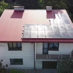 Panel słoneczny o mocy 5,99 kW zainstalowany na czerwonym, blaszanym dachu dwupiętrowego domu z białymi ścianami, widok z góry.