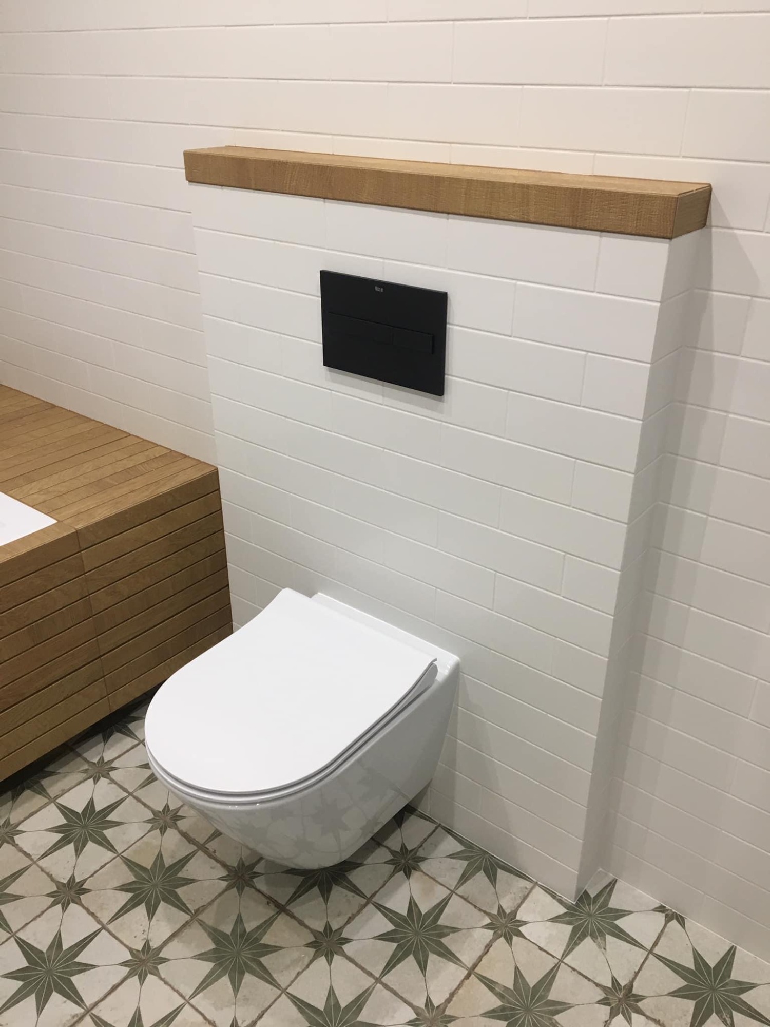 Nowoczesna łazienka z podwieszaną toaletą, czarnym panelem sterującym i drewnianą półką nad nią, białe płytki imitujące cegłę na ścianie, wzorzysta podłoga w gwiazdy.