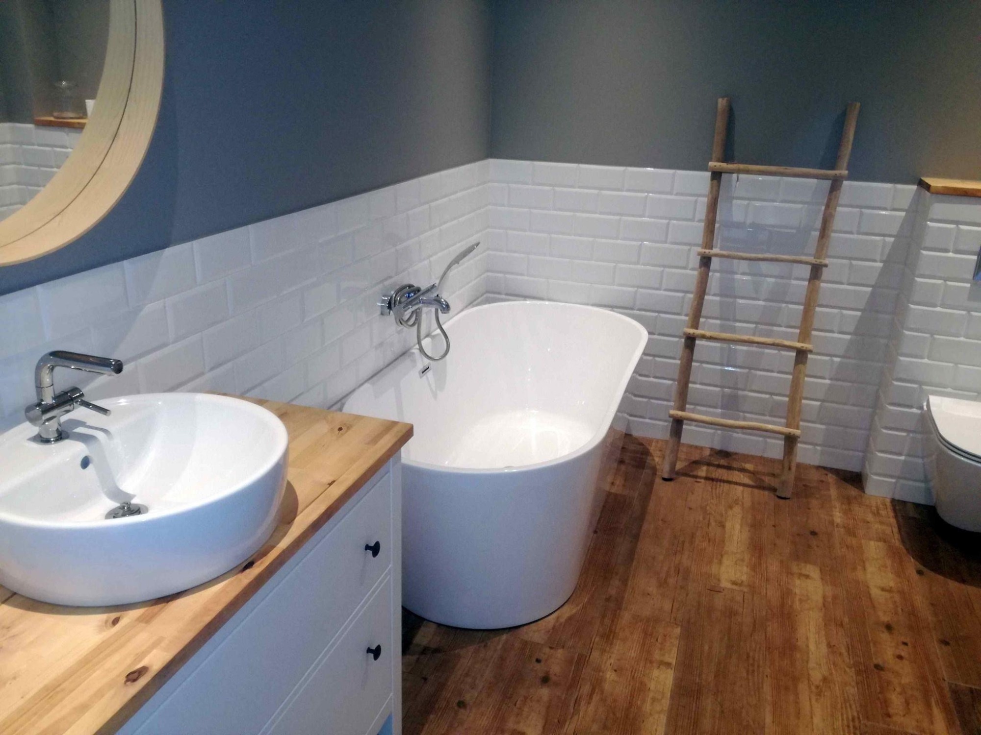 Nowoczesna łazienka z białą wanną wolnostojącą, umywalką nablatową na drewnianym blacie, drewnianą drabinką dekoracyjną i białymi płytkami typu metro na ścianach.