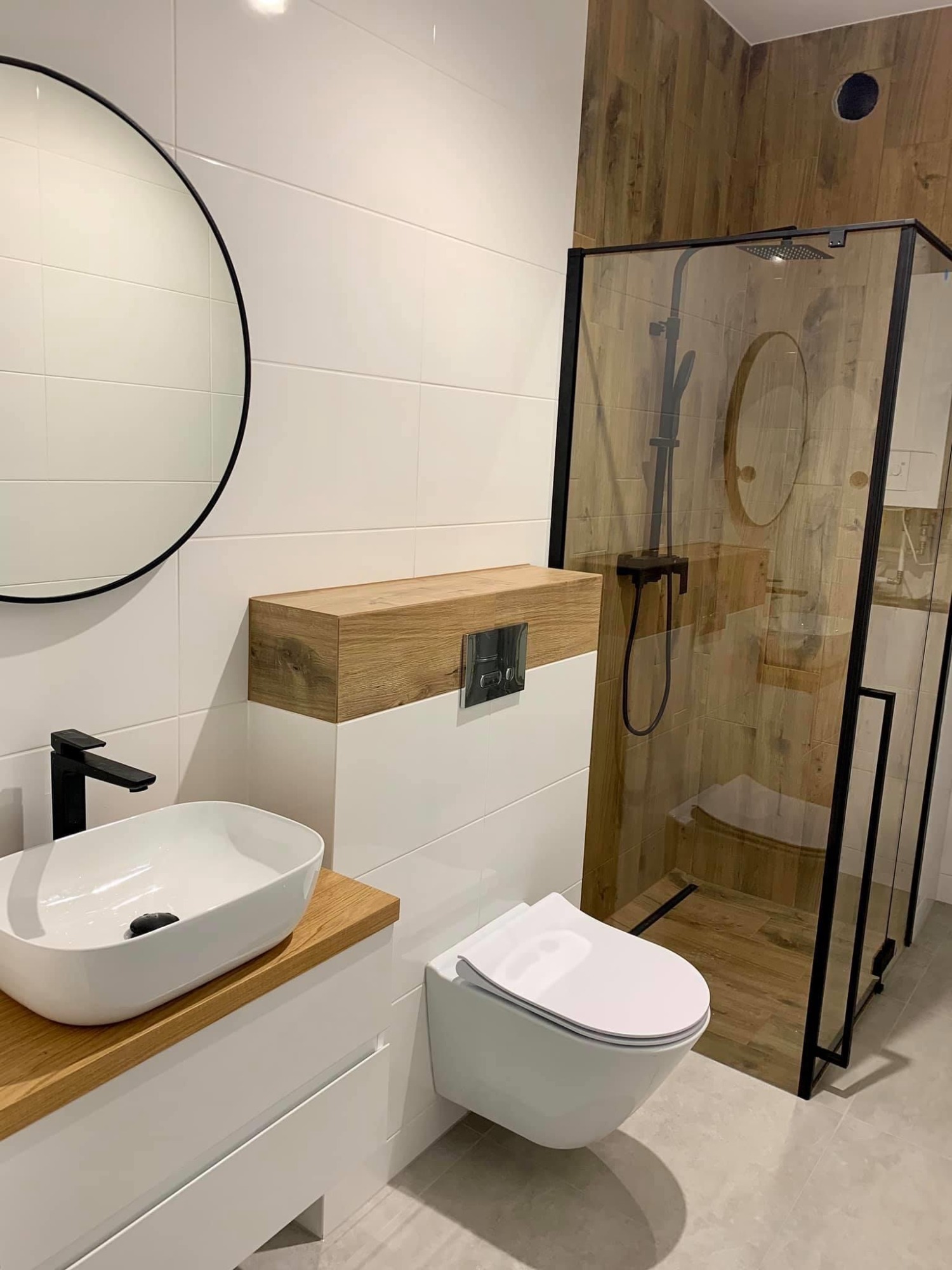 Nowoczesna łazienka z białymi płytkami, drewnianymi akcentami i kabiną prysznicową typu walk-in z czarnymi ramami, okrągłe lustro nad umywalką.
