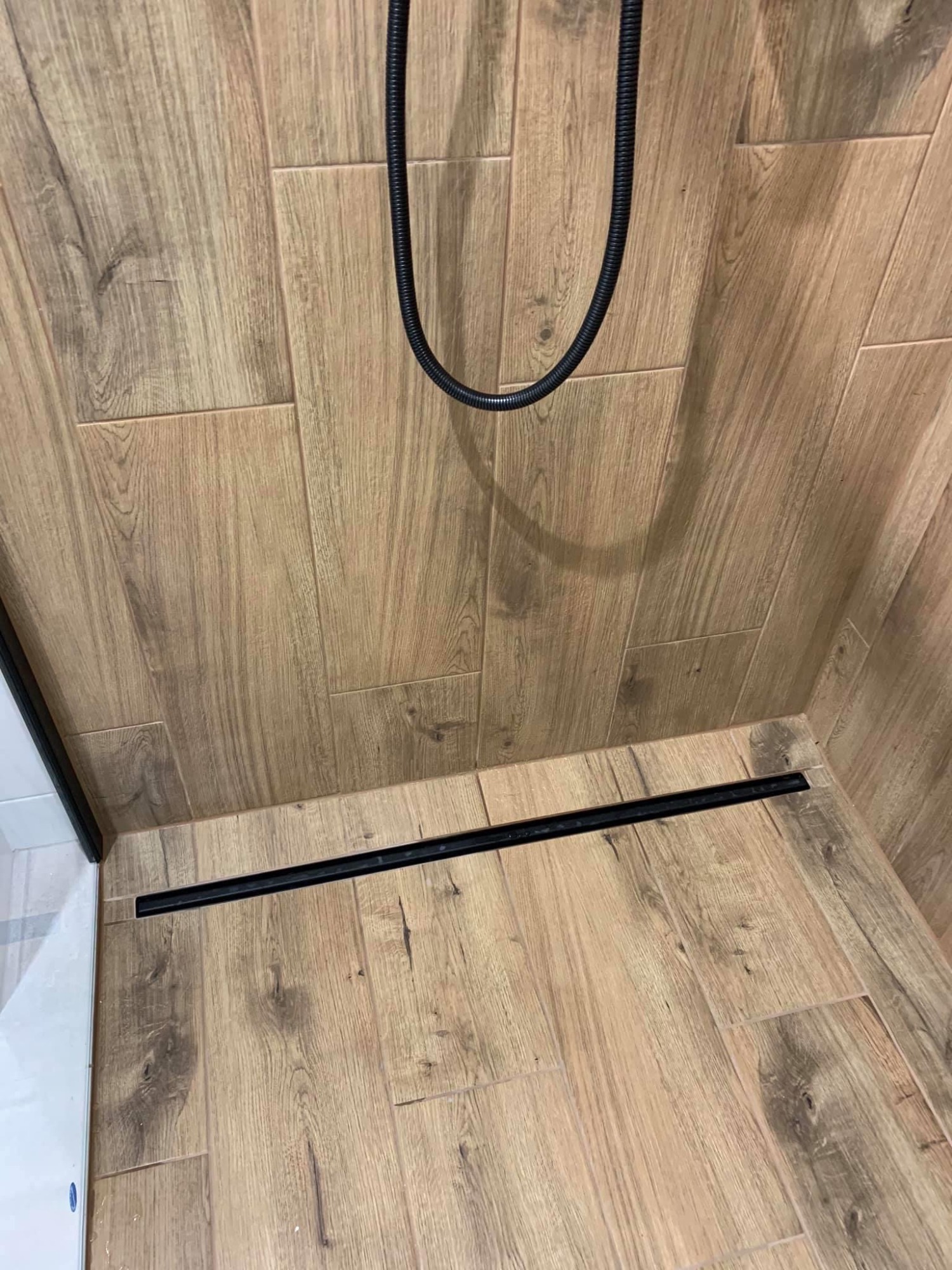 Kabina prysznicowa z odpływem liniowym w podłodze wyłożonej płytkami imitującymi drewno, widoczny fragment węża prysznicowego.