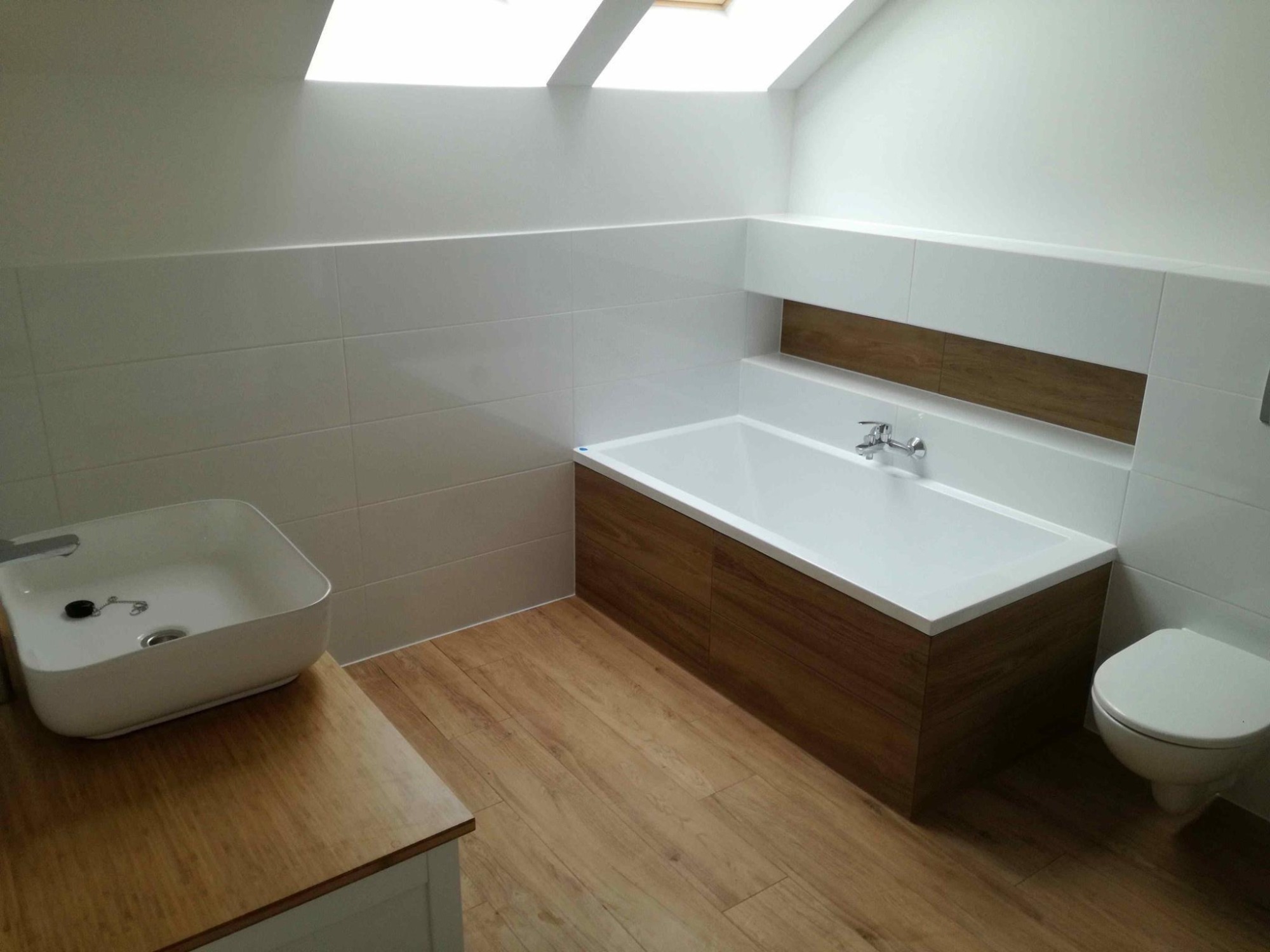 Nowoczesna łazienka na poddaszu z drewnopodobnymi panelami na obudowie wanny i blacie umywalki, białe płytki na ścianach, okno dachowe.