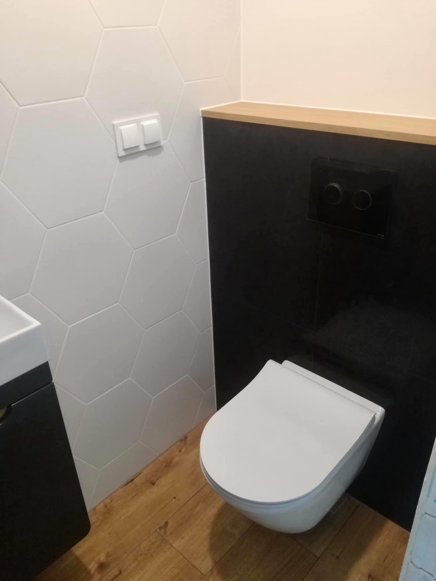 Nowoczesna toaleta podwieszana z czarną ścianą i drewnianym blatem nad nią, heksagonalne białe płytki na ścianie obok, drewnopodobna podłoga.