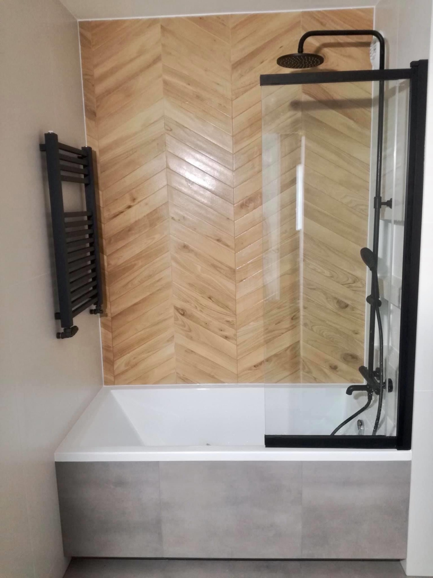 Nowoczesna łazienka z wanną obudowaną szarymi płytkami, drewnopodobną ścianą w jodełkę, czarną armaturą i grzejnikiem drabinkowym.