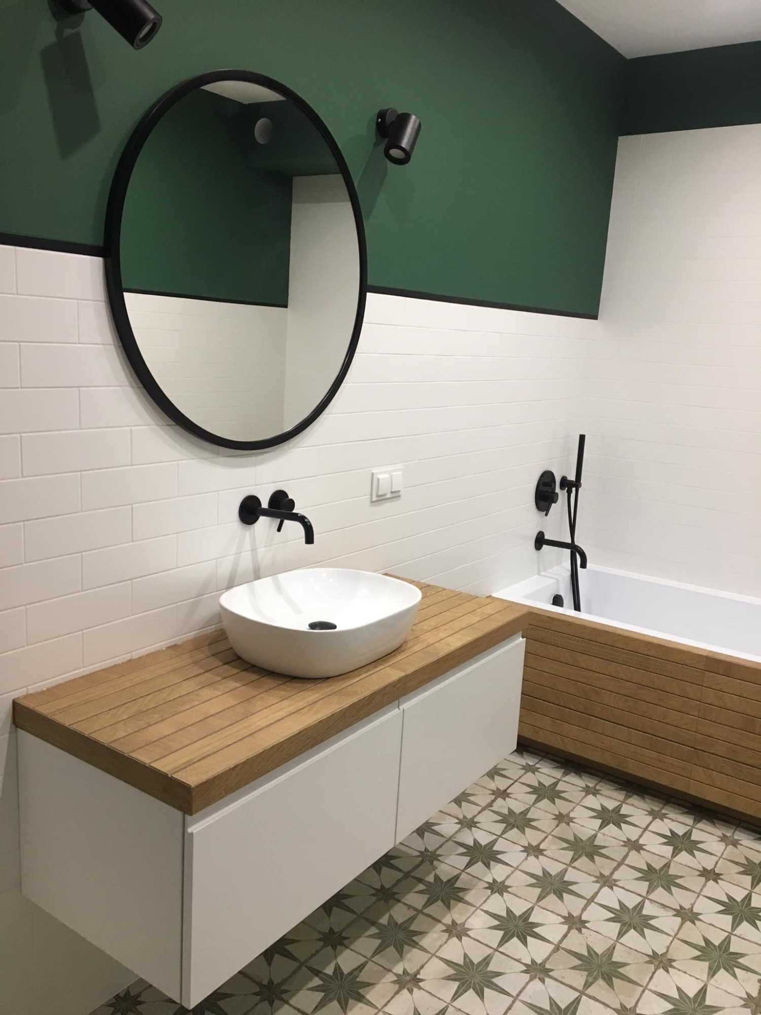 Nowoczesna łazienka z białą armaturą, drewnianym blatem pod umywalką i obudową wanny, zieloną górną częścią ściany i wzorzystą podłogą w stylu retro.