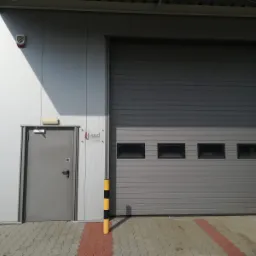 Szary magazyn z bramą segmentową z czterema okienkami, obok wejście z drzwiami, na ścianie logo firmy SOD i czujnik alarmowy, na podłodze kostka brukowa.