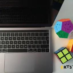 Laptop z otwartym ekranem prezentującym kod, obok ułożone kostki Rubika o różnych kształtach i kolorach, na białym tle, z widocznym logo firmy w prawym dolnym rogu.