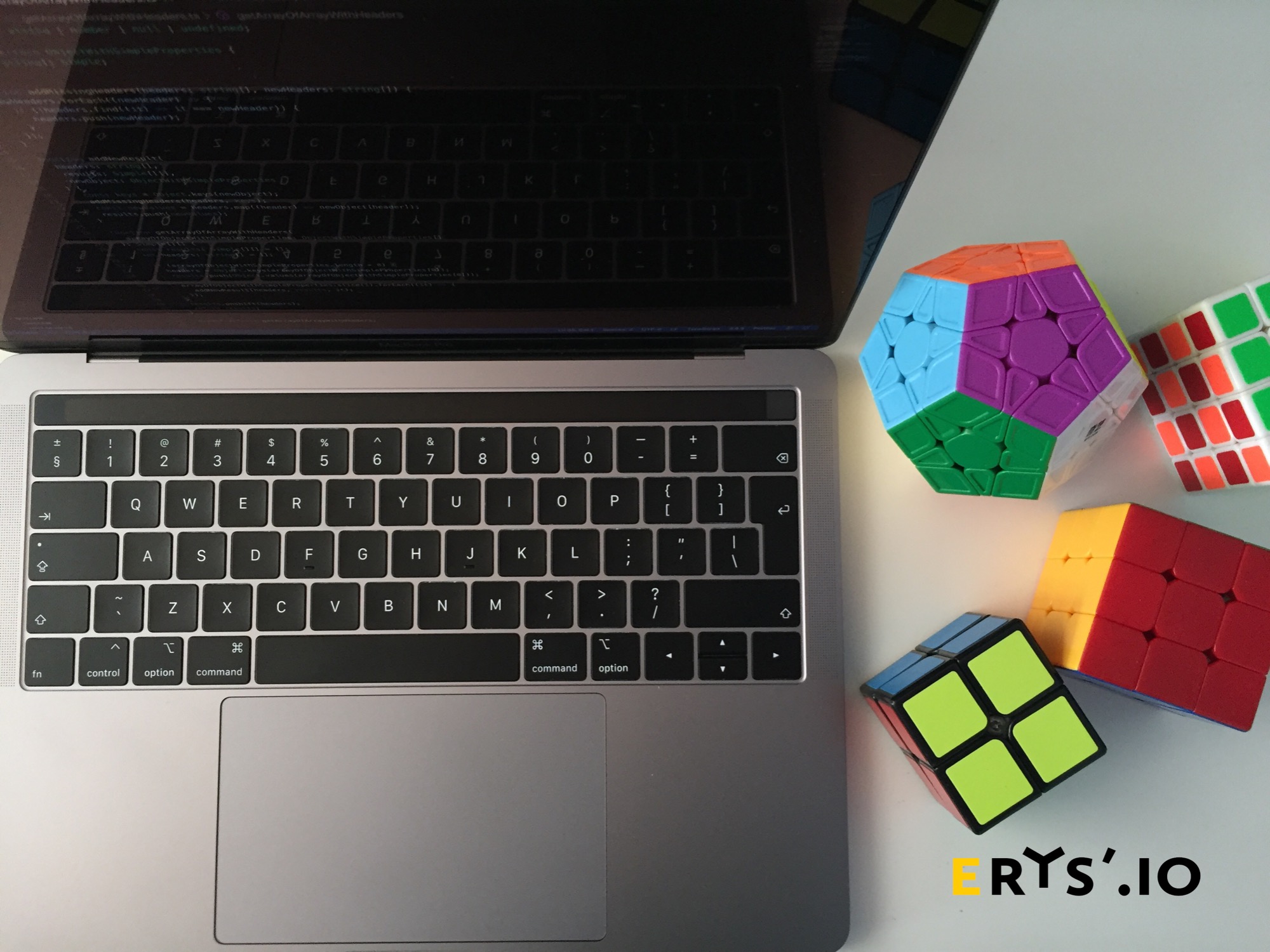 Laptop z otwartym ekranem prezentującym kod, obok ułożone kostki Rubika o różnych kształtach i kolorach, na białym tle, z widocznym logo firmy w prawym dolnym rogu.