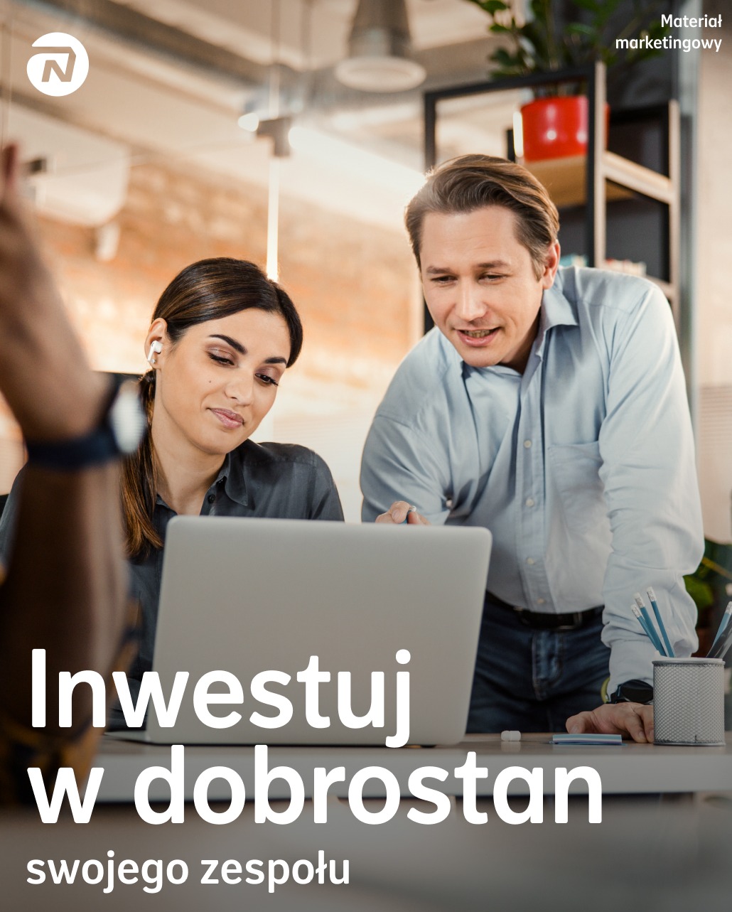 Dwie osoby pochylają się nad laptopem w biurze. Tekst: Inwestuj w dobrostan swojego zespołu. Materiał marketingowy. Koncepcja ubezpieczeń grupowych.