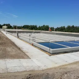 Budowa oczyszczalni ścieków z widocznymi zbiornikami retencyjnymi wypełnionymi wodą, placem budowy i koparką w tle pod błękitnym niebem.