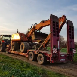 Pomarańczowa koparka kołowa transportowana na niskopodwoziowej, czerwonej przyczepie ciągniętej przez czarny ciągnik rolniczy na polnej drodze o zmierzchu.