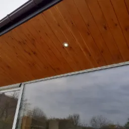 Drewniana podbitka dachowa z zamontowanym oświetleniem punktowym, widoczny fragment okna z odbiciem krajobrazu.