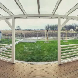 Zadaszony taras z brązową podłogą, białą konstrukcją i widokiem na ogród z domkiem dla dzieci w oddali.