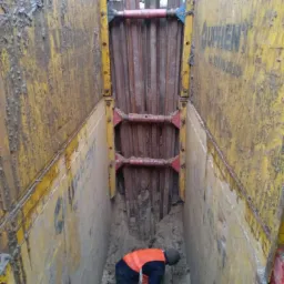 Głęboki wykop zabezpieczony żółtymi ścianami oporowymi z widocznym pracownikiem w pomarańczowej kamizelce, wykonującym prace na dnie wykopu.