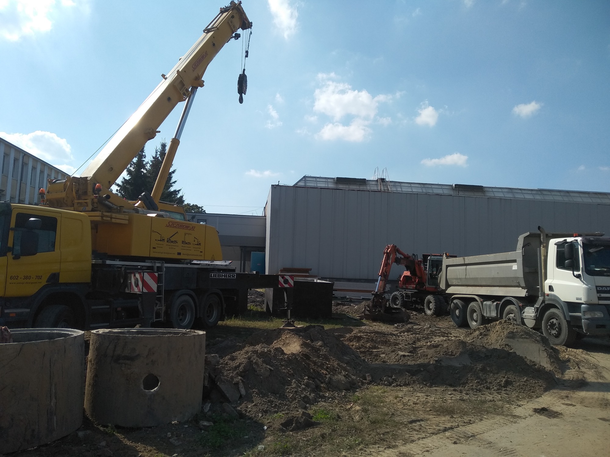 Żółty dźwig Liebherr, koparka i wywrotka na placu budowy; w pierwszym planie betonowe kręgi z otworami, prawdopodobnie do budowy kanalizacji.