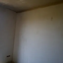 Wnętrze pomieszczenia po nałożeniu tynków; widoczne gładkie, jasnoszare ściany, surowy stan wykończenia, trzy gniazdka elektryczne w ścianie, oraz fragment okna z widokiem na zieleń na zewnątrz.