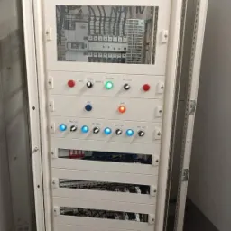 Rozdzielnica elektryczna w obudowie metalowej, z widocznymi przewodami, bezpiecznikami i panelami kontrolnymi z diodami LED w kolorach czerwonym, zielonym i niebieskim.