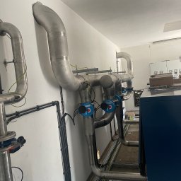 Paweł Kowalski P.K.-SPAW - Instalacja przemysłowa z rurami ze stali nierdzewnej, wyposażona w zawory z niebieskimi pokrętłami, prowadzona wzdłuż ściany wewnątrz budynku.