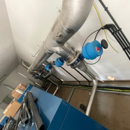 Paweł Kowalski P.K.-SPAW - Instalacja przemysłowa z rur ze stali nierdzewnej z niebieskimi zaworami, prowadząca pod sufitem w pomieszczeniu technicznym, widoczne kable elektryczne i fragment niebieskiej obudowy urządzenia.