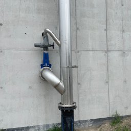 Paweł Kowalski P.K.-SPAW - Metalowa rura pionowa z zaworem i kolankiem przymocowana do betonowej ściany, widoczne ślady po szalunkach, trawa u podstawy.