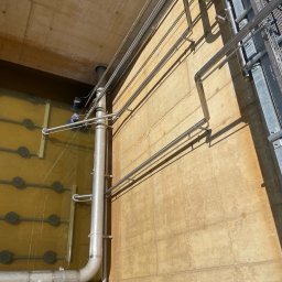 Paweł Kowalski P.K.-SPAW - Instalacja rur ze stali nierdzewnej wewnątrz betonowego zbiornika z wodą, widoczna drabina i pracownik w wodzie w tle.