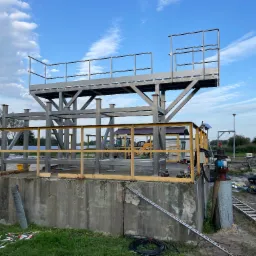 Konstrukcja stalowa platformy z barierkami, częściowo zmontowana na betonowym fundamencie, na tle błękitnego nieba z chmurami i krajobrazu z drzewami i budynkami w oddali, z widocznym sprzętem...