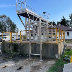 Paweł Kowalski P.K.-SPAW - Metalowa konstrukcja platformy z drabinką i żółtymi barierkami na betonowym fundamencie, na tle budynku i błękitnego nieba.