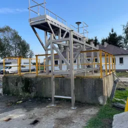 Metalowa konstrukcja platformy z drabinką i żółtymi barierkami na betonowym fundamencie, na tle budynku i błękitnego nieba.