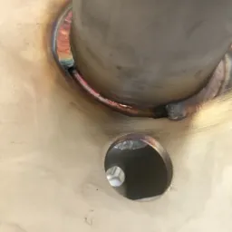 Wspawana rura w metalową płytę, widoczne ślady spawania z charakterystycznymi przebarwieniami termicznymi i otw&oacute;r w płycie z wykończeniem krawędzi.