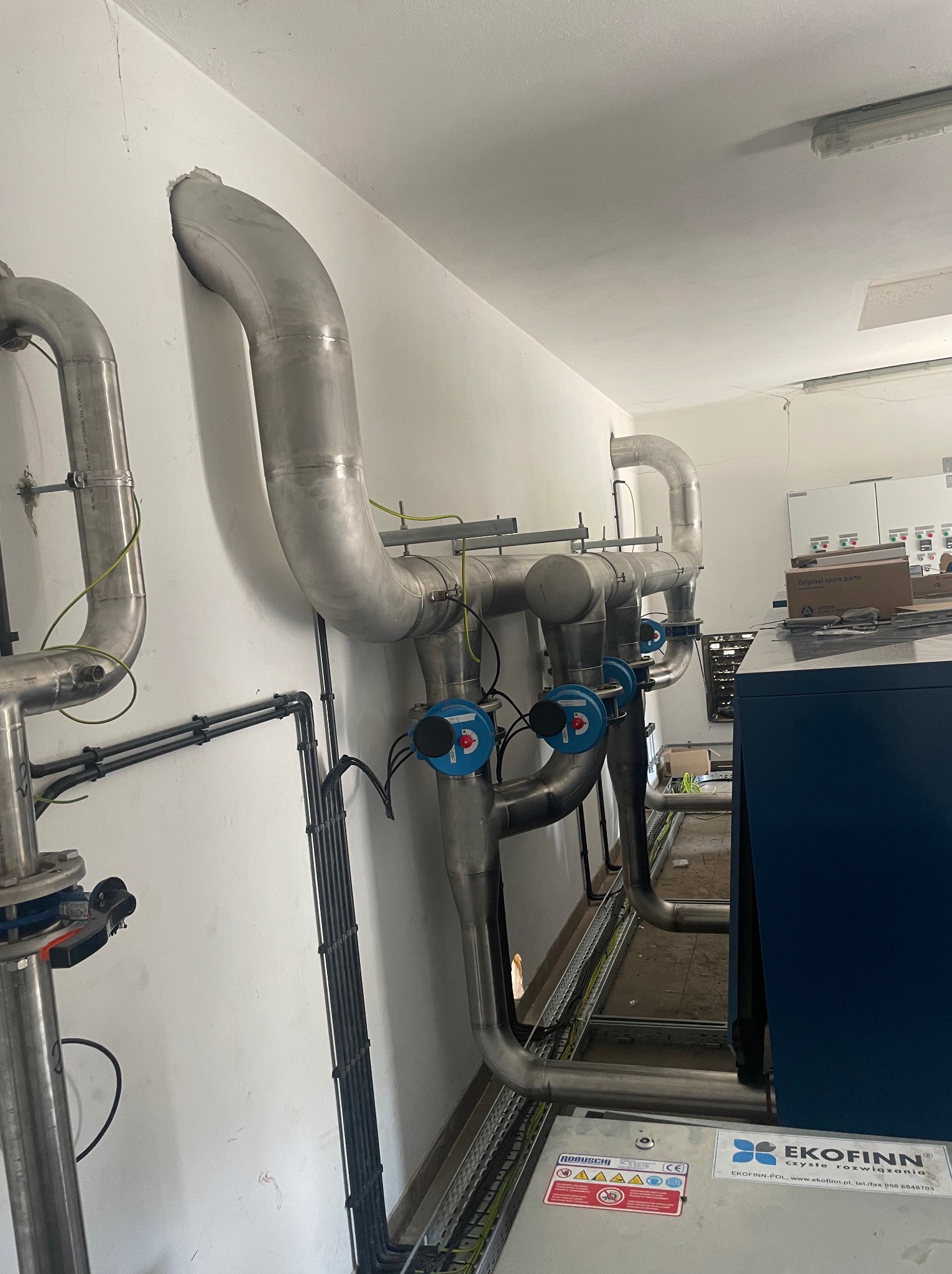 Instalacja przemysłowa z rurami ze stali nierdzewnej, wyposażona w zawory z niebieskimi pokrętłami, prowadzona wzdłuż ściany wewnątrz budynku.