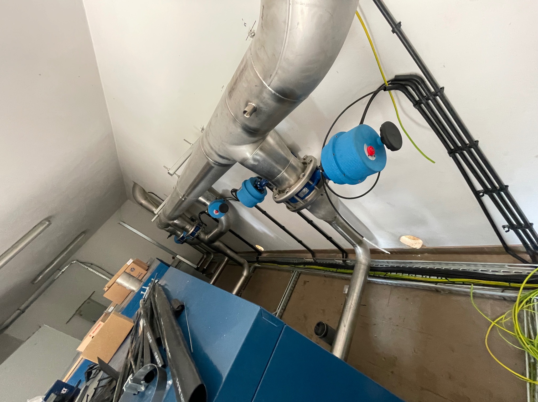 Instalacja przemysłowa z rur ze stali nierdzewnej z niebieskimi zaworami, prowadząca pod sufitem w pomieszczeniu technicznym, widoczne kable elektryczne i fragment niebieskiej obudowy urządzenia.