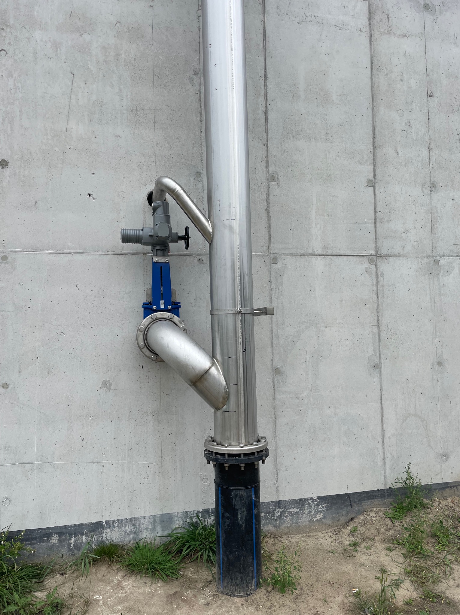 Metalowa rura pionowa z zaworem i kolankiem przymocowana do betonowej ściany, widoczne ślady po szalunkach, trawa u podstawy.