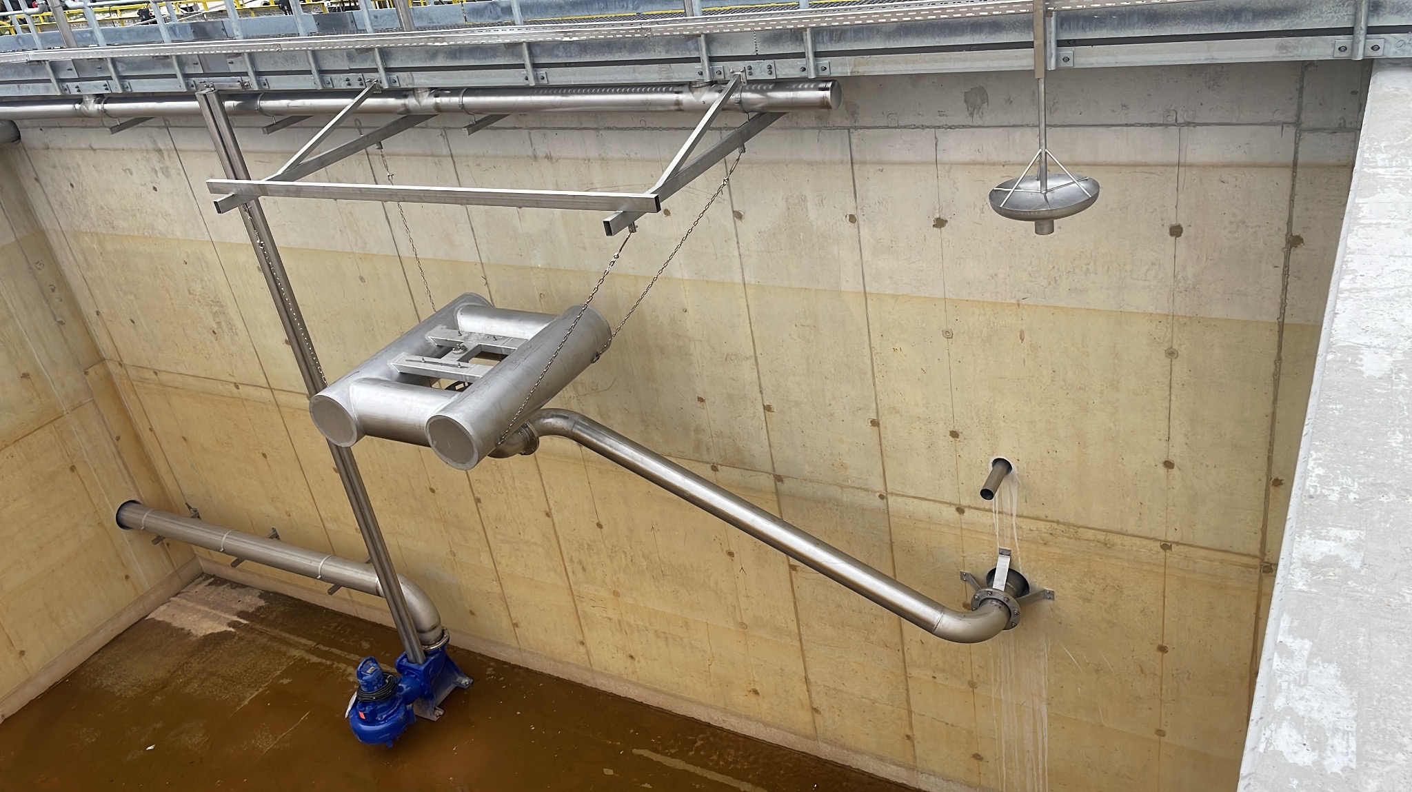 Instalacja przemysłowa z rurami i pompą w betonowym zbiorniku, widoczne elementy konstrukcji stalowej podwieszone na łańcuchach, ściekająca woda.