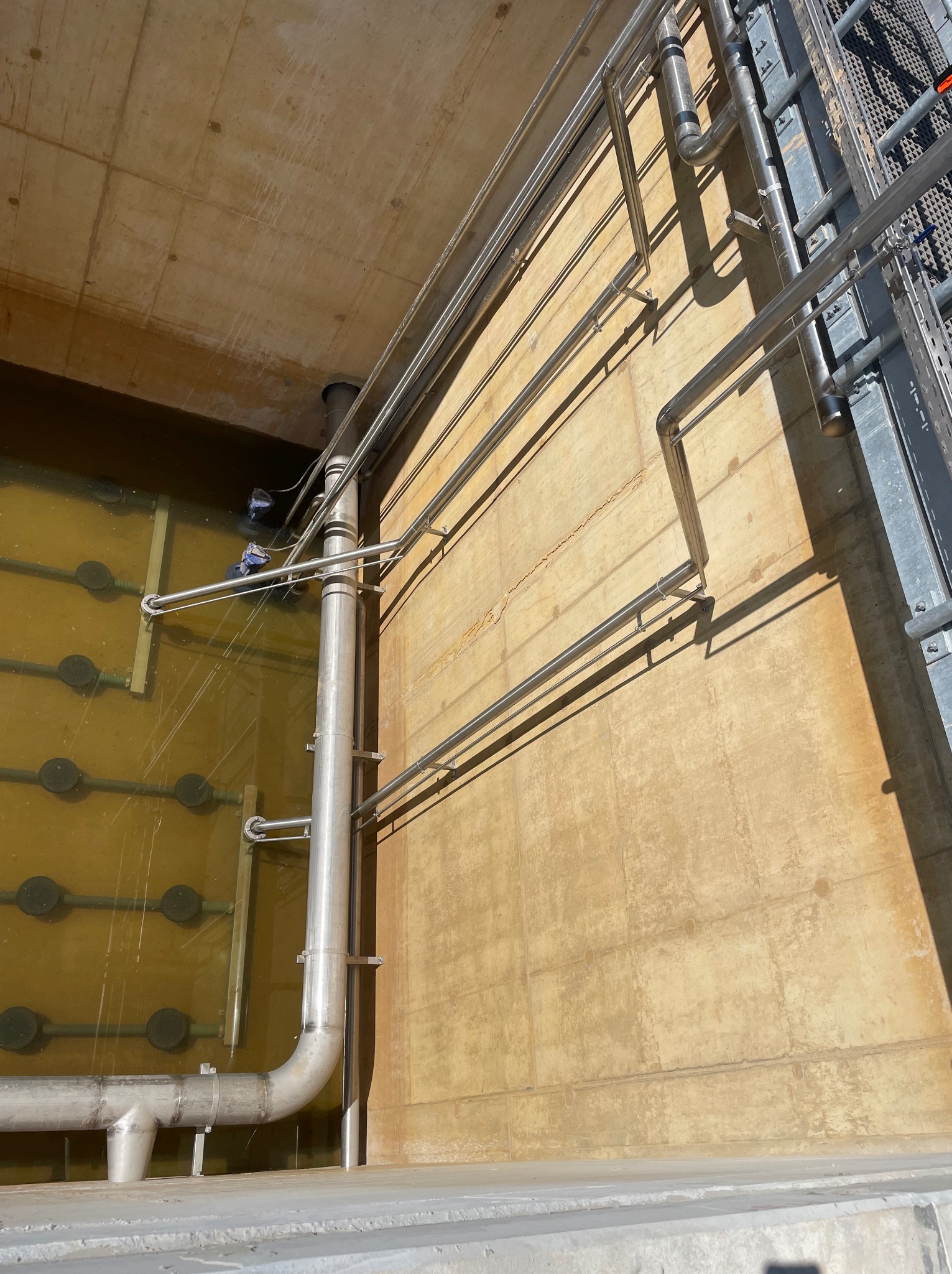 Instalacja rur ze stali nierdzewnej wewnątrz betonowego zbiornika z wodą, widoczna drabina i pracownik w wodzie w tle.