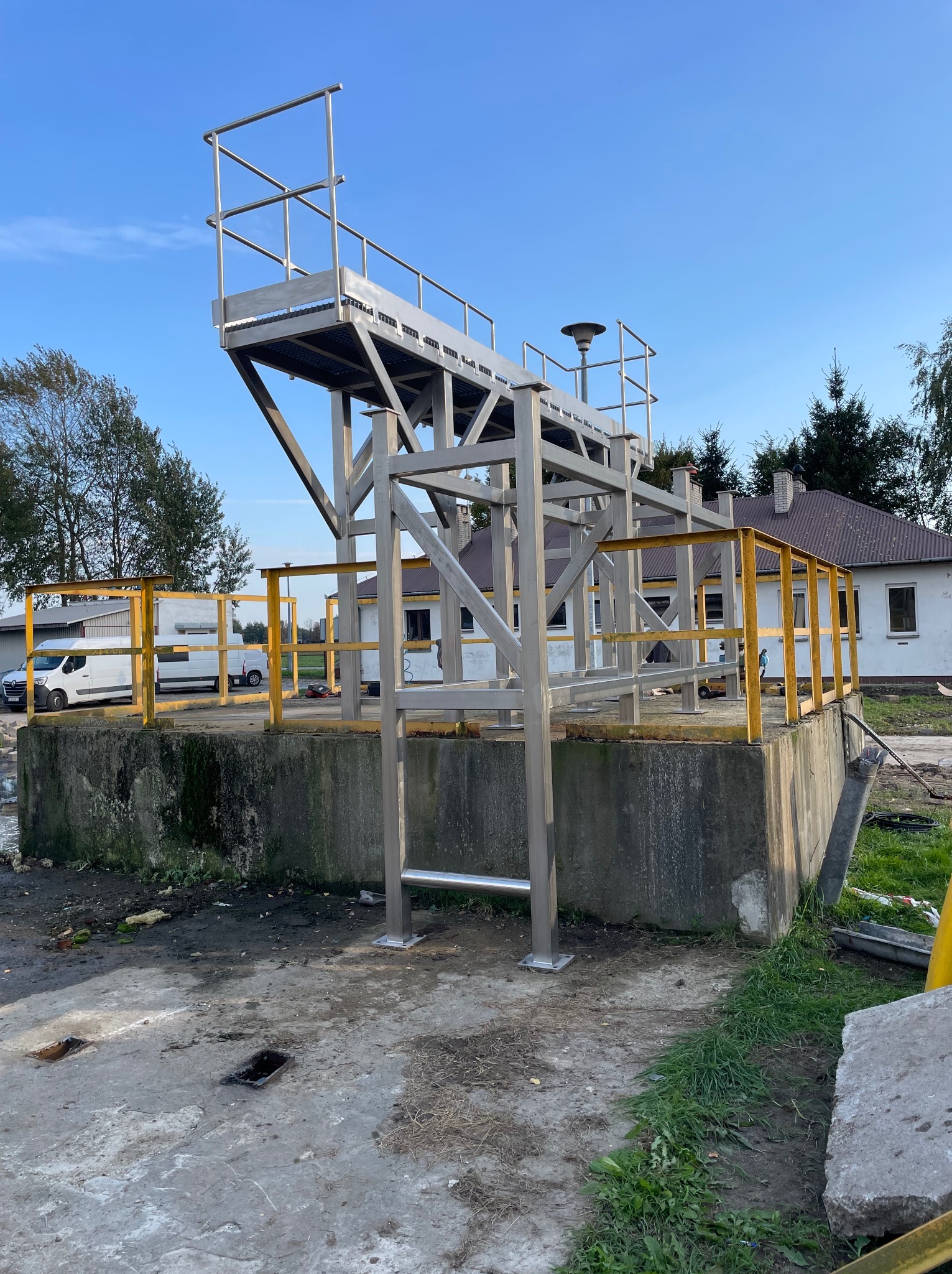 Metalowa konstrukcja platformy z drabinką i żółtymi barierkami na betonowym fundamencie, na tle budynku i błękitnego nieba.